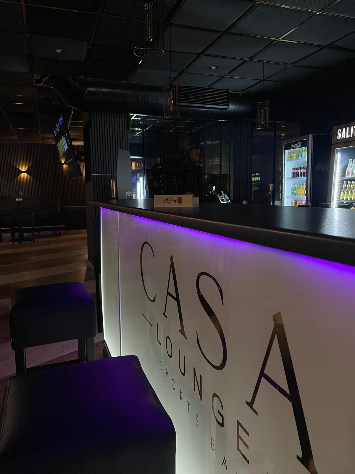 Die beleuchtete Bar der Casa Lounge mit Casa-Lounge-Schriftzug und violettem LED-Licht